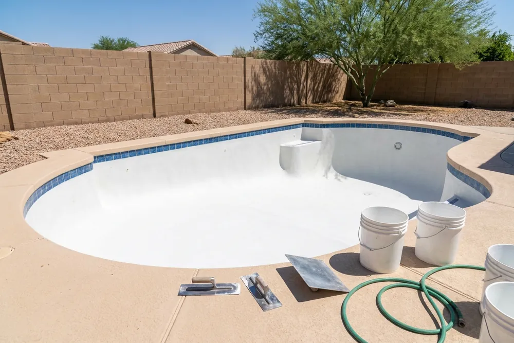 Drained backyard pool with fresh white plaster, finishing trowels, buckets and a hose on the deck, and a block wall and palo verde tree behind